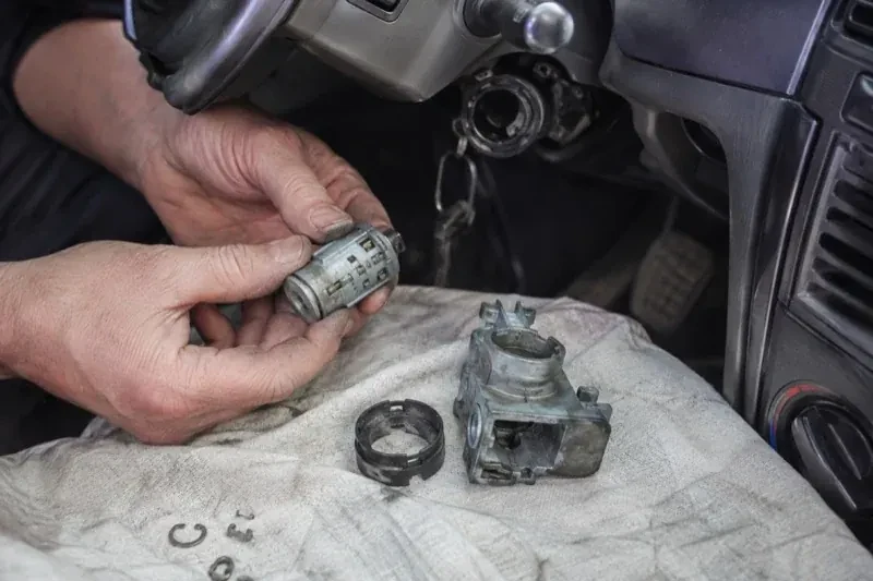 Hands holding a removed ignition cylinder beside a car steering column, with the housing and small parts on a cloth. — Ignition Lock Repair, Brampton